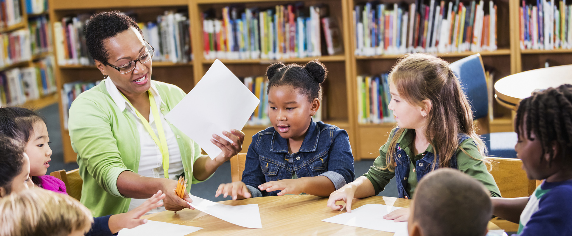 A teacher sitting at a round table handing out paper and pencils to elementary students. 