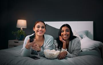Two girls laying in bed watching TV with a bowl of popcorn.
