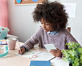 An African American girl sitting at a desk holding a mobile phone while writing notes 