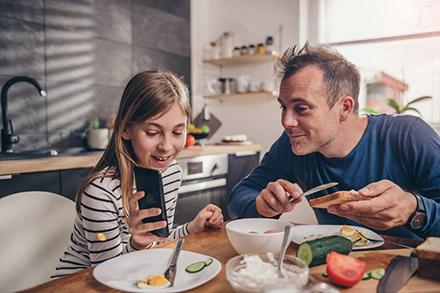 Father and daughter sitting at a kitchen table looking at a smart phone while eating