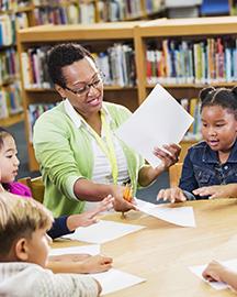 A teacher sitting at a round table handing out paper and pencils to elementary students.