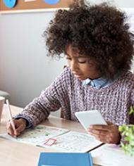 An African American girl sitting at a desk holding a mobile phone while writing notes 
